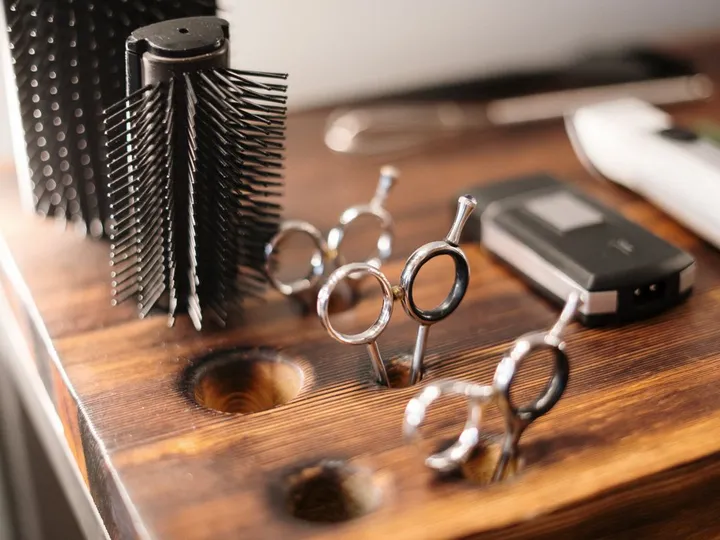 Brosse ronde, trois paires de ciseaux de coupe et une tondeuse posées sur la tablette de bois d’un poste de coiffure.